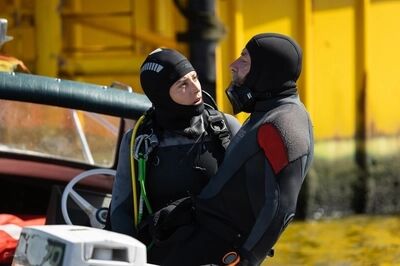 Duikers in wetsuits staan dicht bij elkaar naast een boot, met een gele achtergrond van een gebouw.